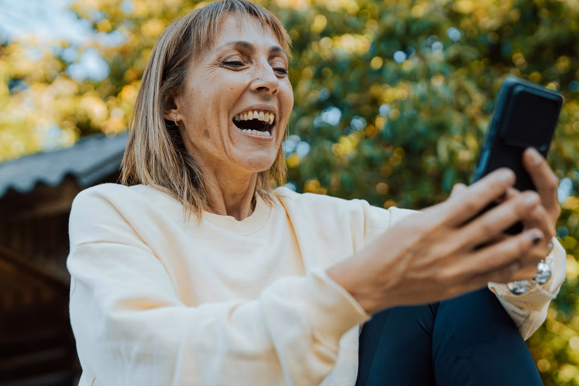 Smiling woman outdoors holding a smartphone, appearing to take a selfie or video call.