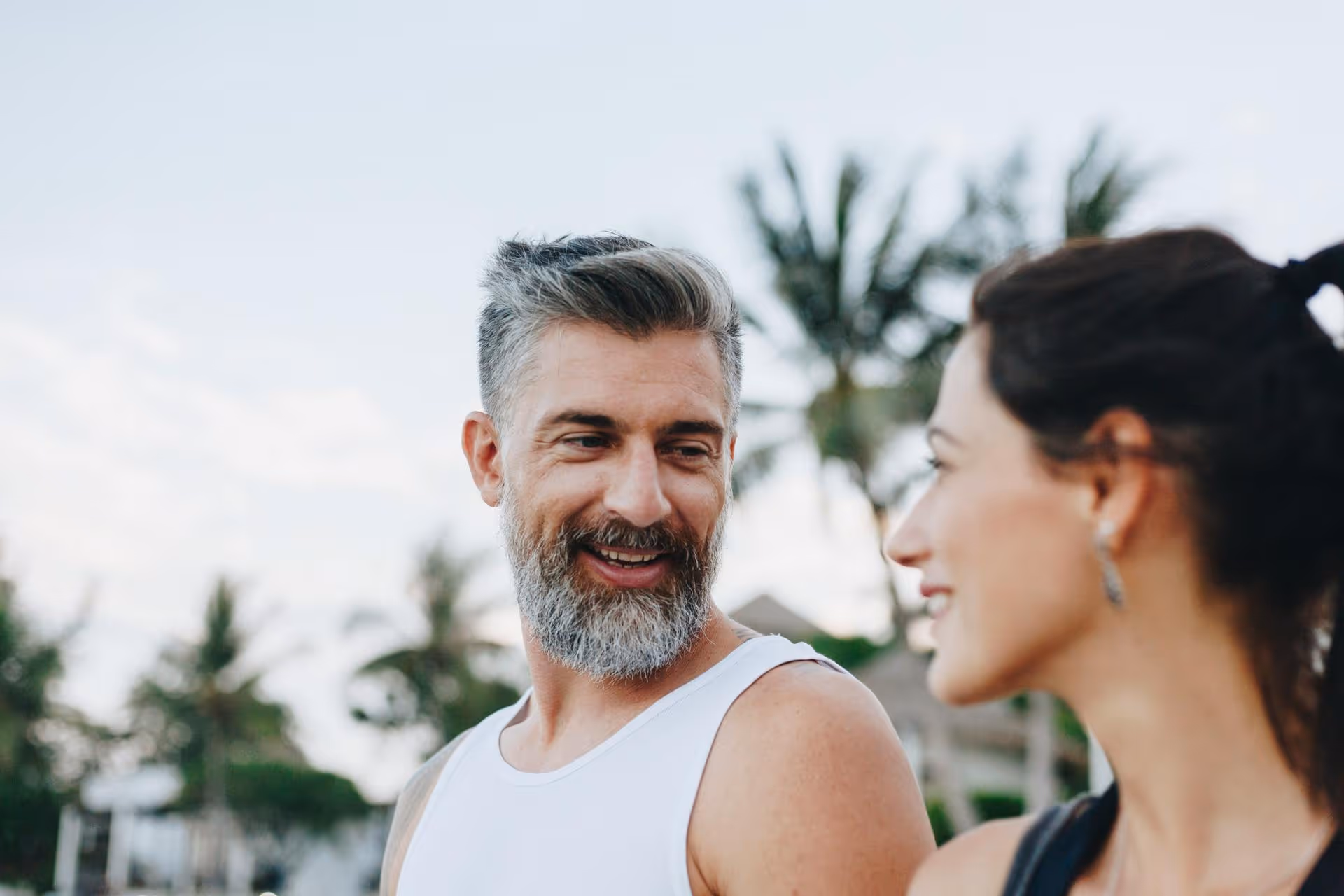 Smiling man and woman looking at each other outdoors with palm trees in the background.