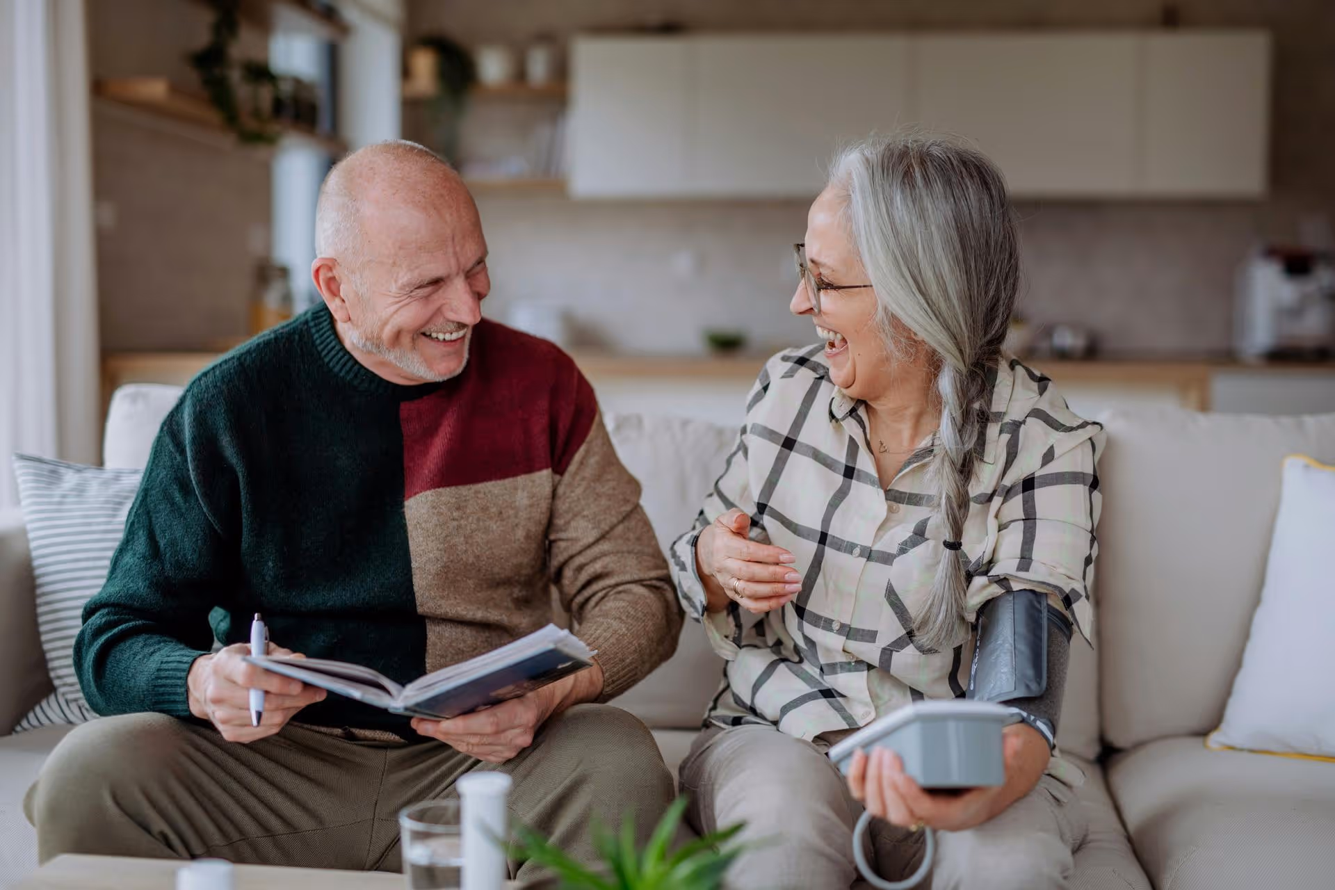 Elderly couple sitting on a couch, the woman measuring her blood pressure while both laugh together.