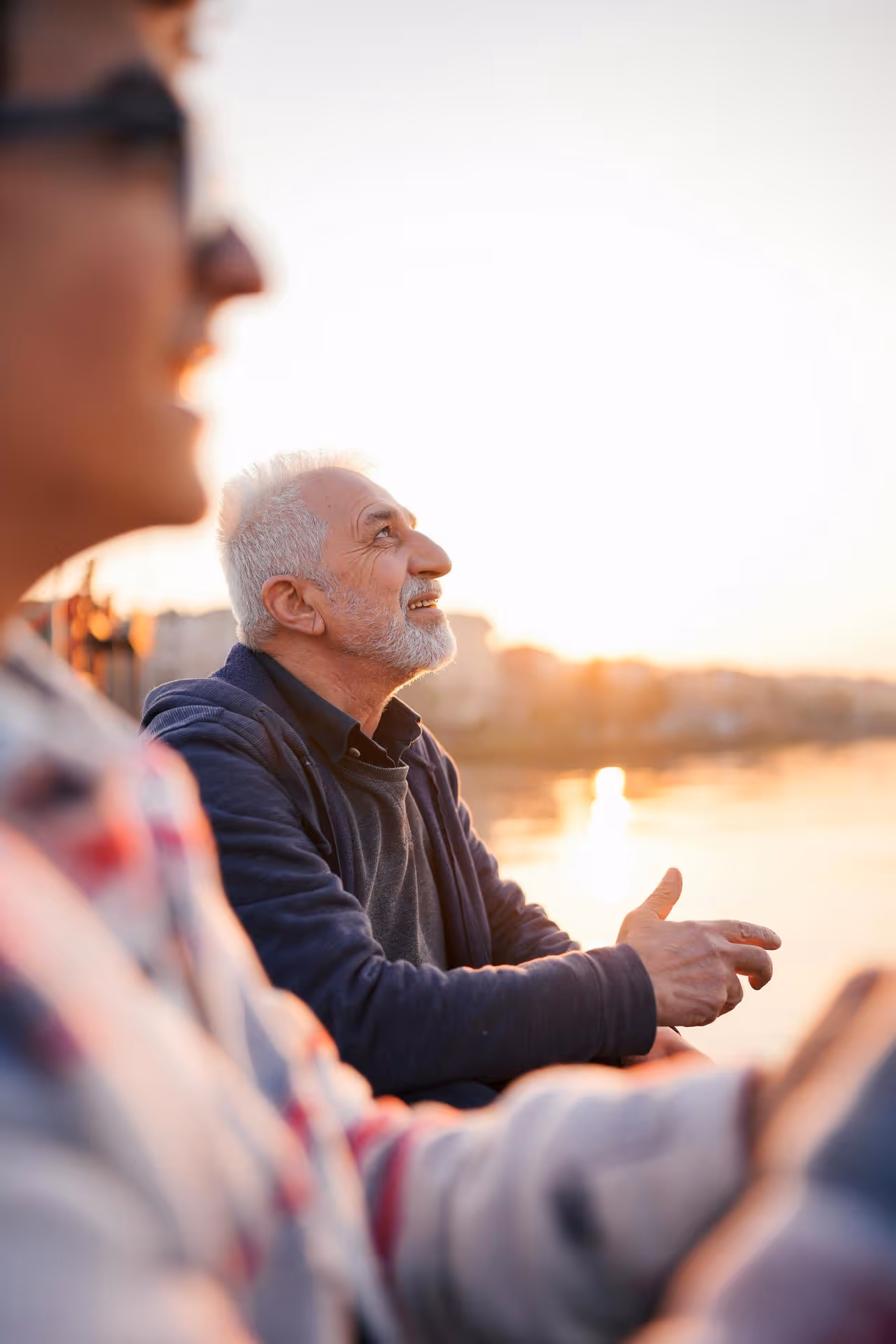 Elderly man with gray hair and beard looking up and smiling, sitting outdoors at sunset with a blurred person in the foreground.