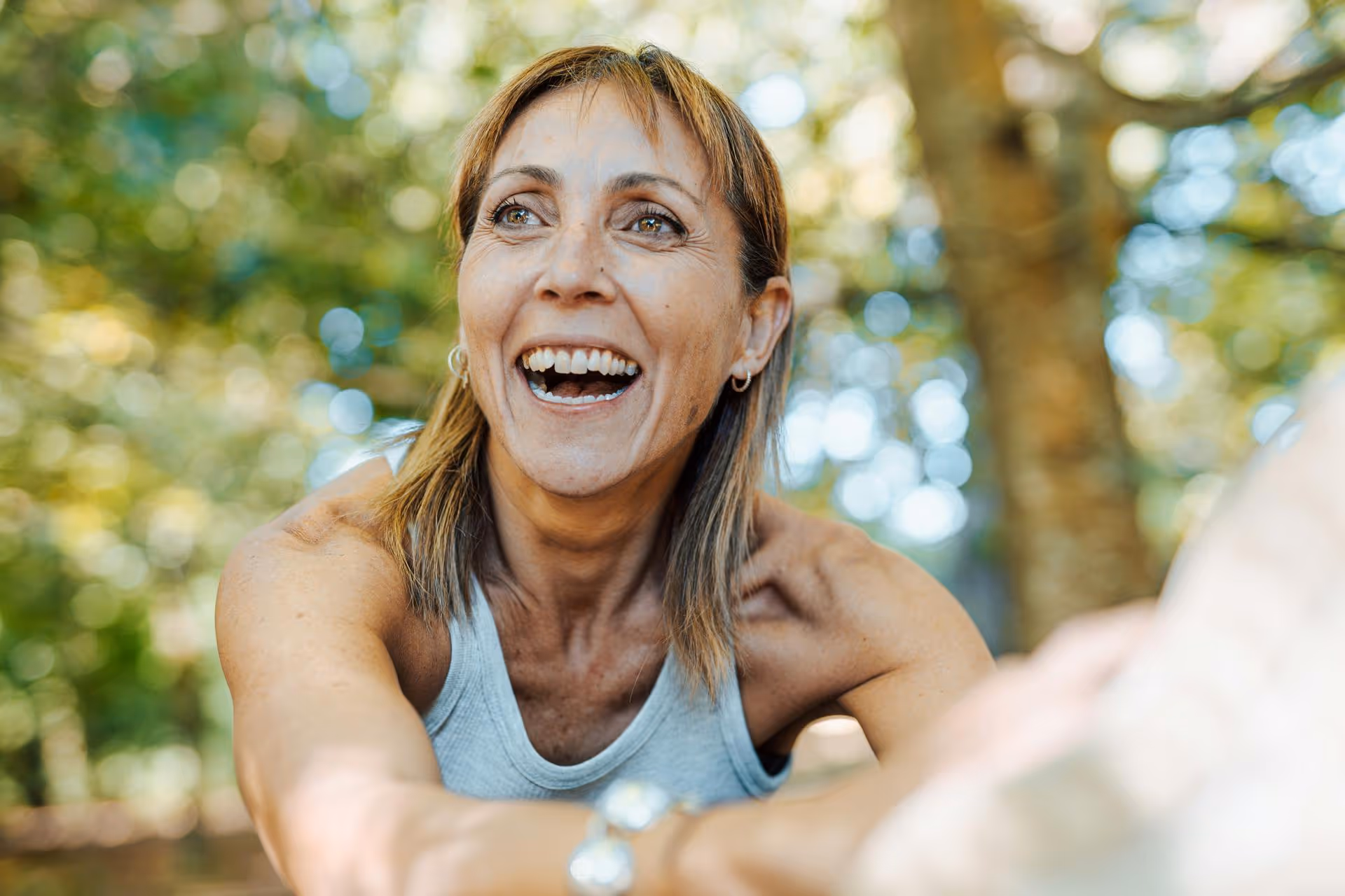Smiling woman with light brown hair and silver hoop earrings reaching out in an outdoor setting with blurred trees in the background.