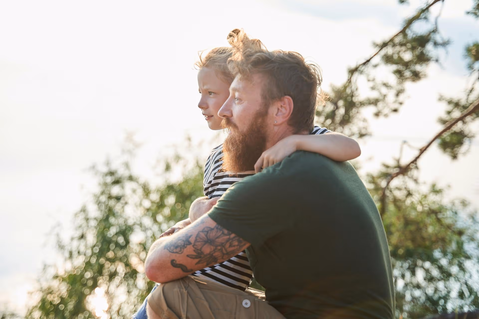 Bearded man with tattoos carrying a young child on his back outdoors during daylight.