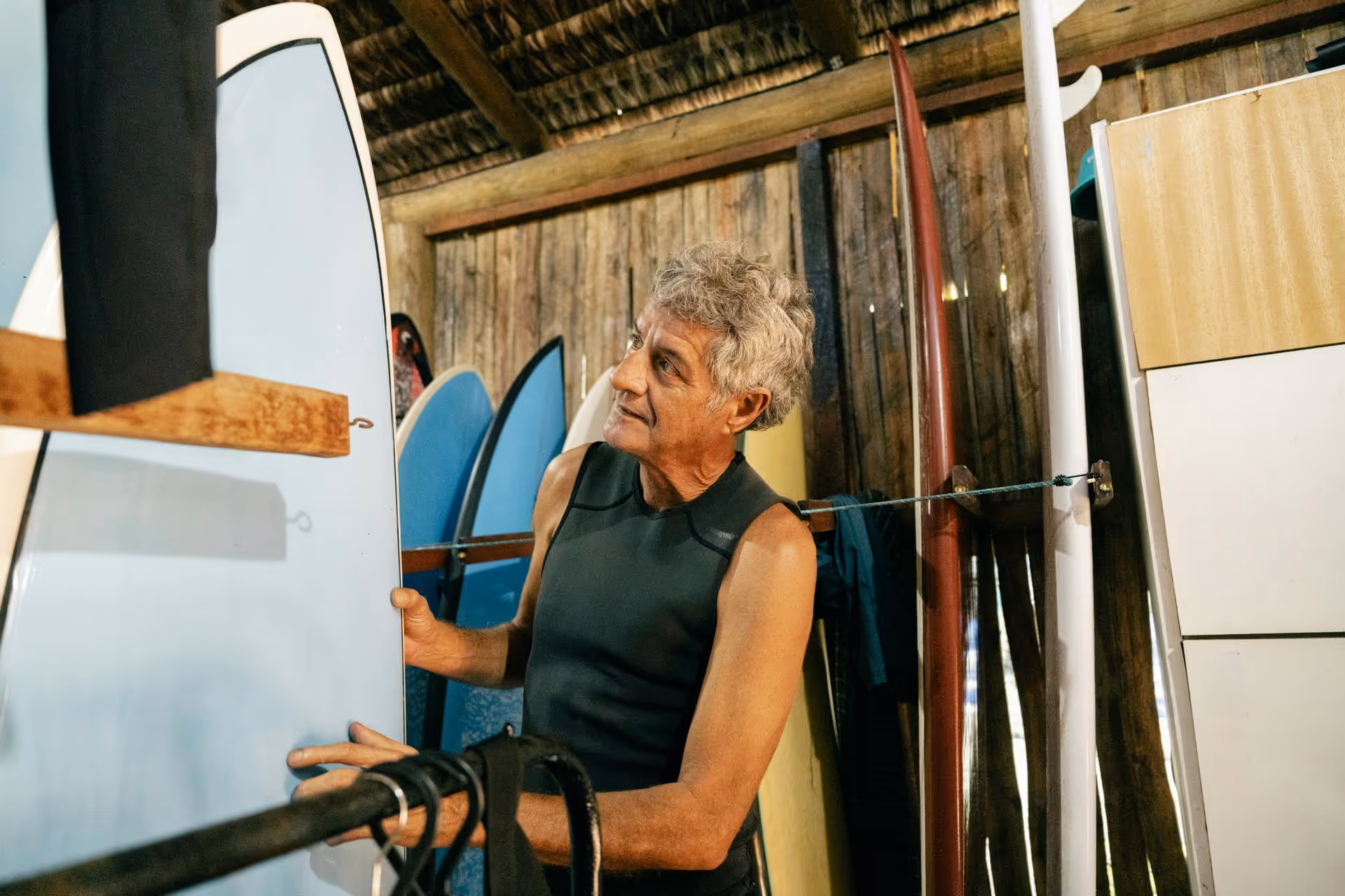 Man in wetsuit inspecting a surfboard in a wooden surfboard storage room with multiple boards.