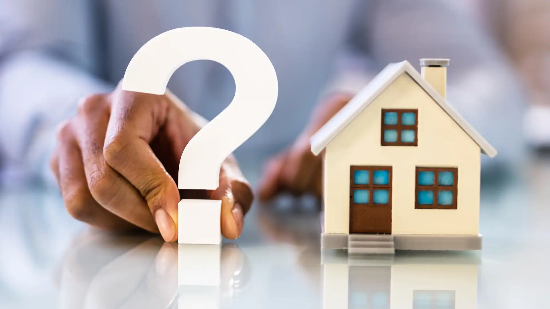 a person holding a question mark between their index finger and thumb with a house figurine on the right all with tones of blue
