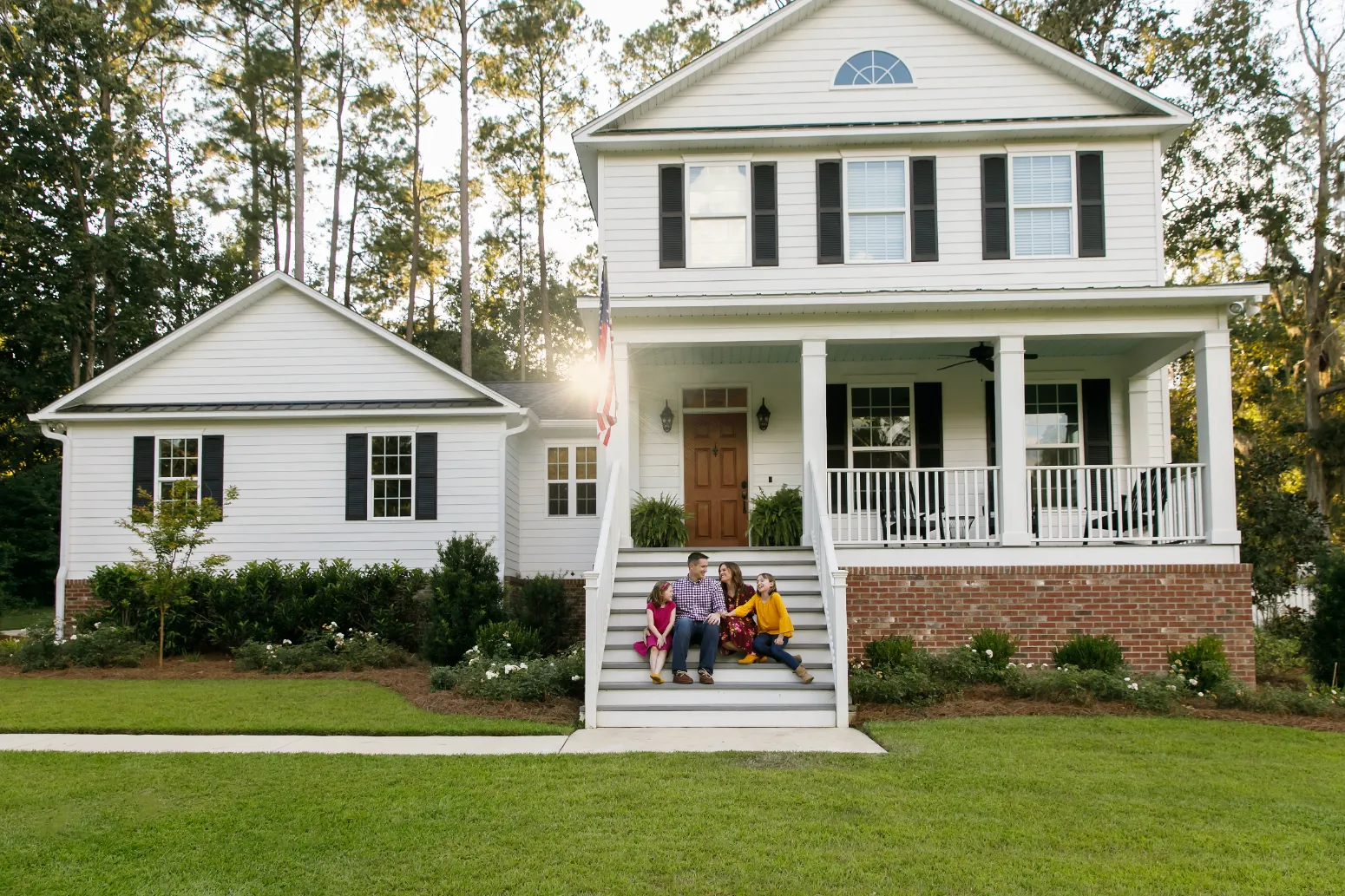 a white 2 story home with a staircase leading up to the front door where a family of 4 are sitting on the stairs