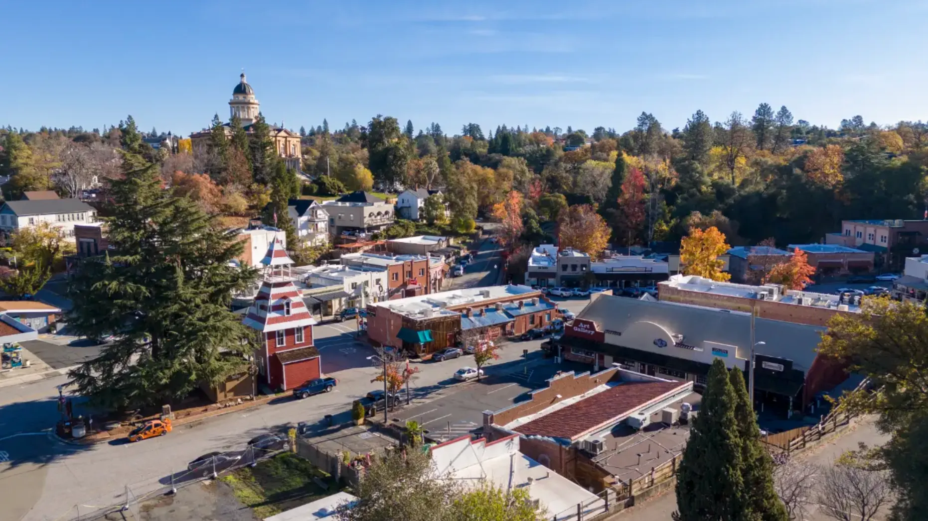 An aerial shot shows a town with historic charm, autumn trees, a standout red and white pointed building, and a domed structure in the distance under a clear blue sky.