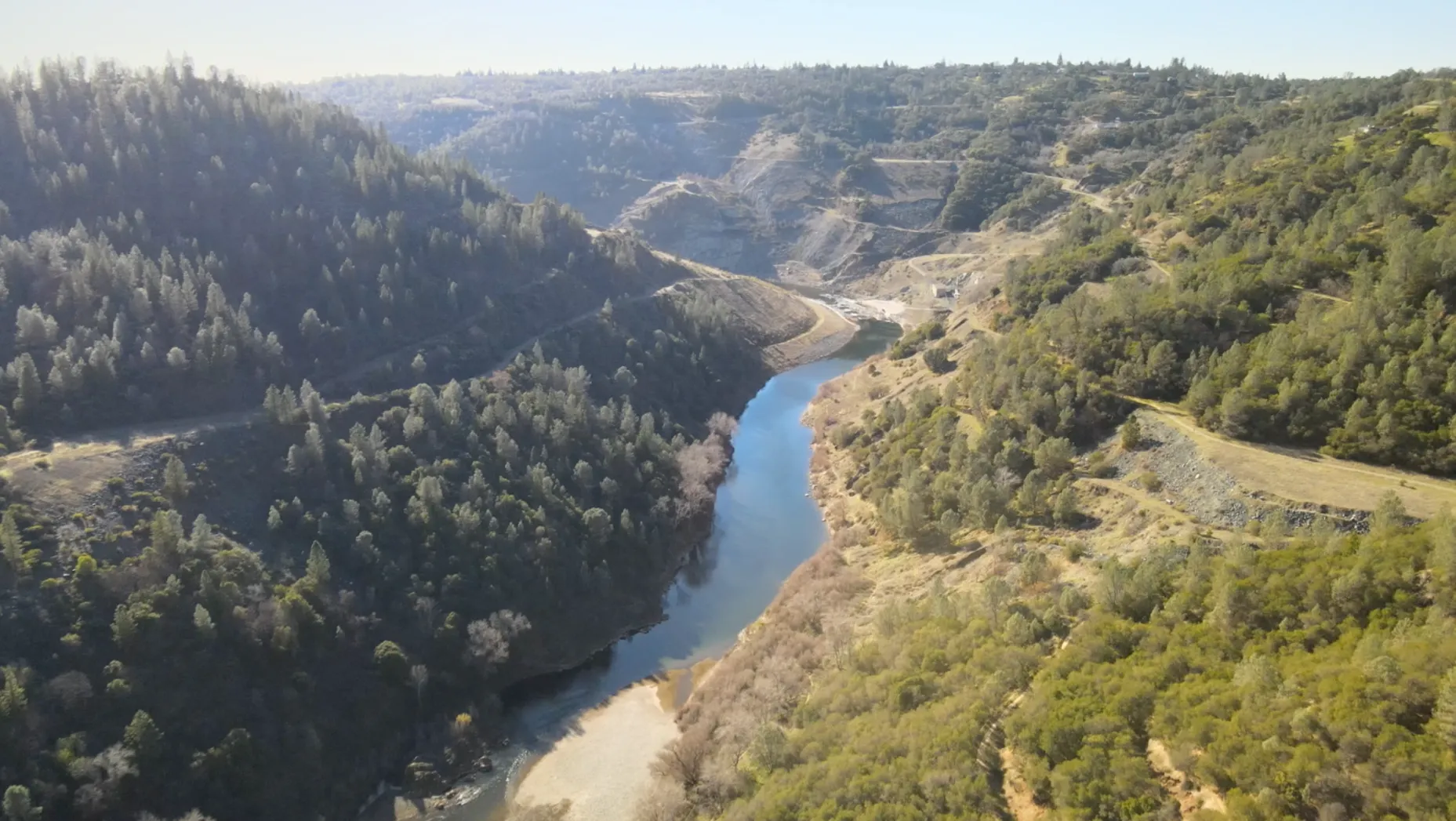  An aerial view captures a serpentine river cutting through a verdant valley with rolling hills. Sparse vegetation clings to the sunlit slopes, contrasting the dense, shadowy tree cover. The landscape exudes tranquility and natural beauty.