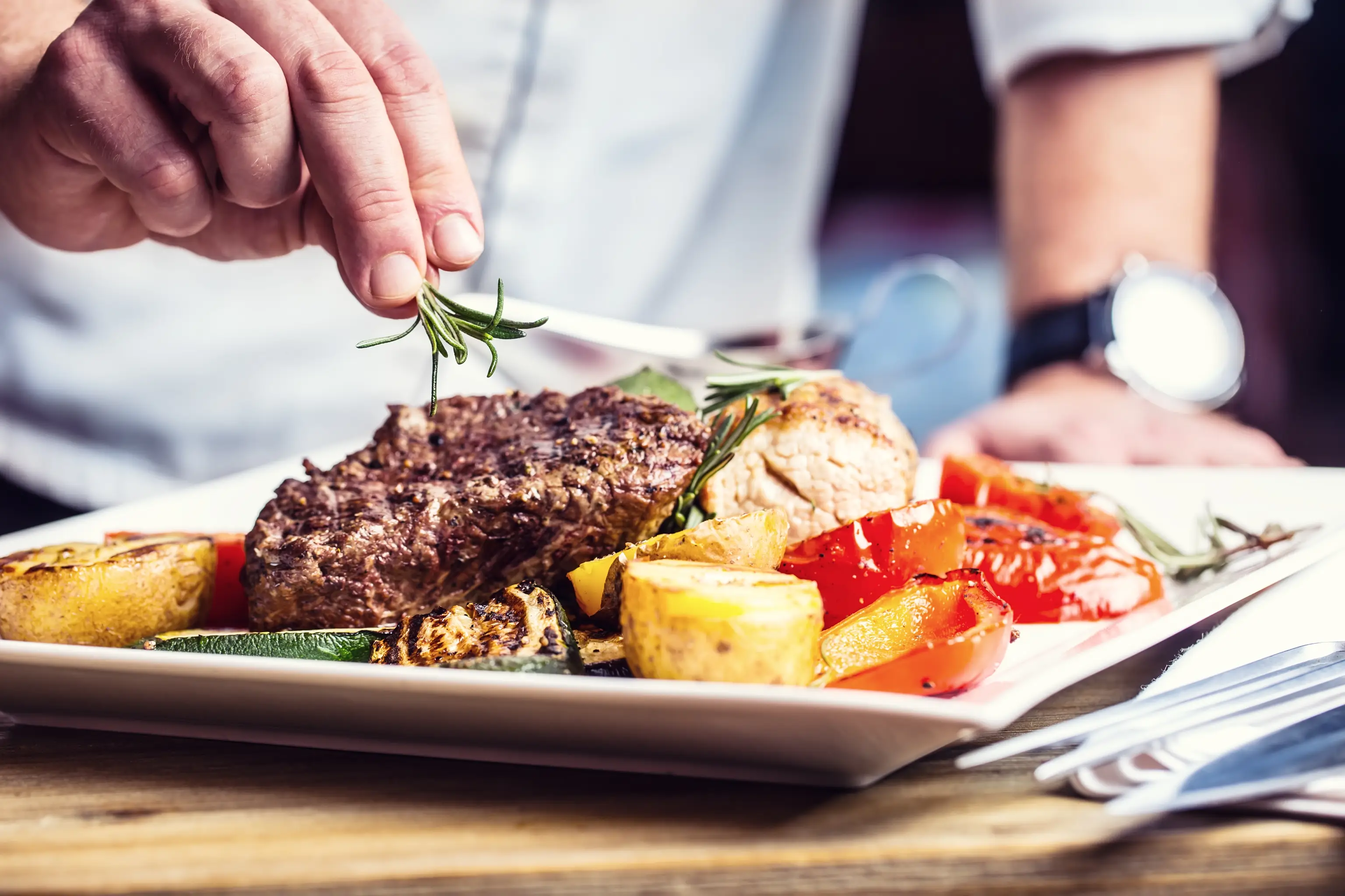 A chef garnishes a plate with fresh rosemary. The dish includes a juicy, grilled steak, roasted potatoes, and colorful grilled vegetables such as red bell peppers and zucchini, presented on a white rectangular plate. The setting is a rustic wooden table.