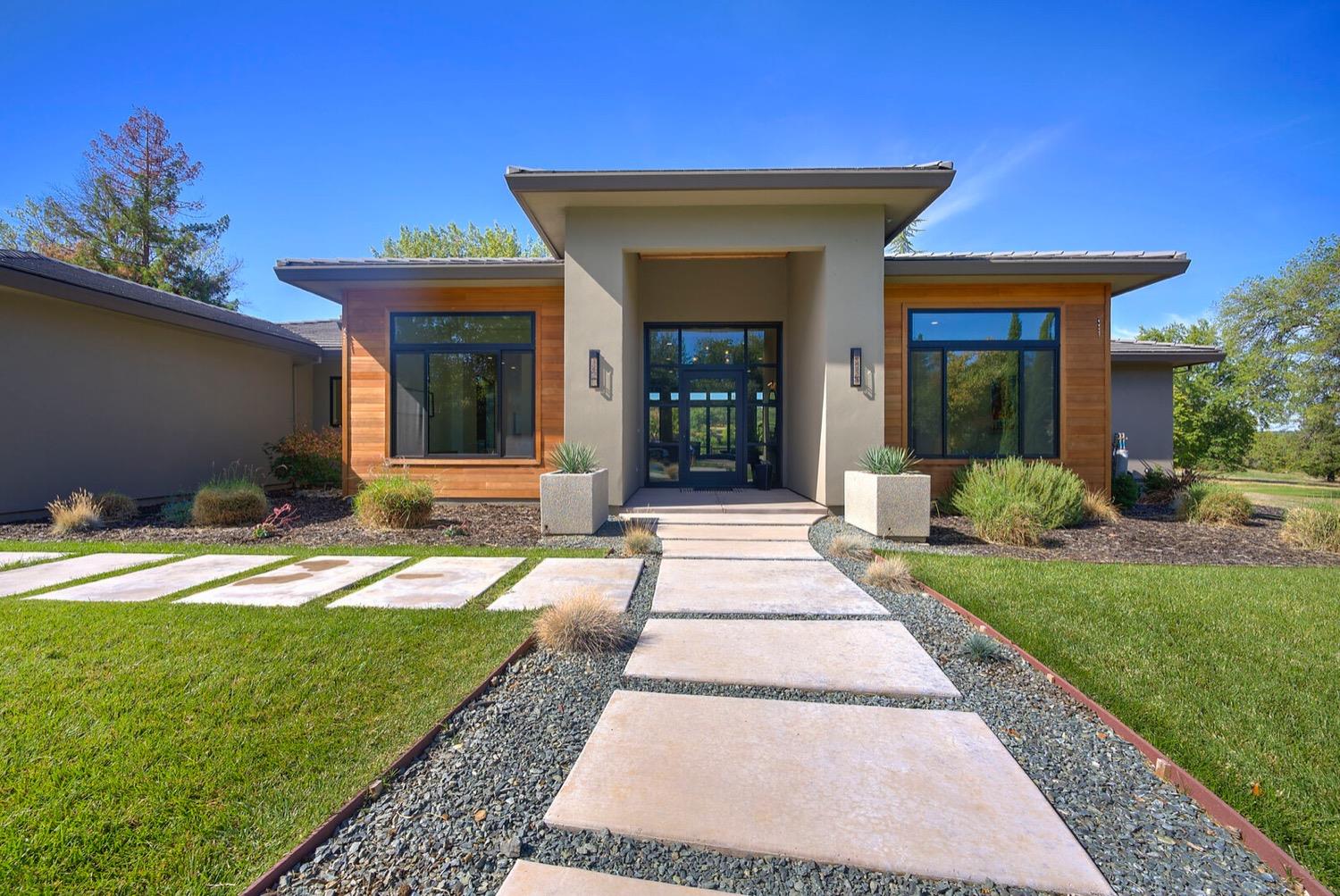 A modern home in Auburn, CA, featuring a sleek, contemporary design with large windows, a minimalist front pathway, and well-maintained landscaping. The home is set against a bright blue sky on a sunny day, highlighting the clean lines and natural elements of the architecture.