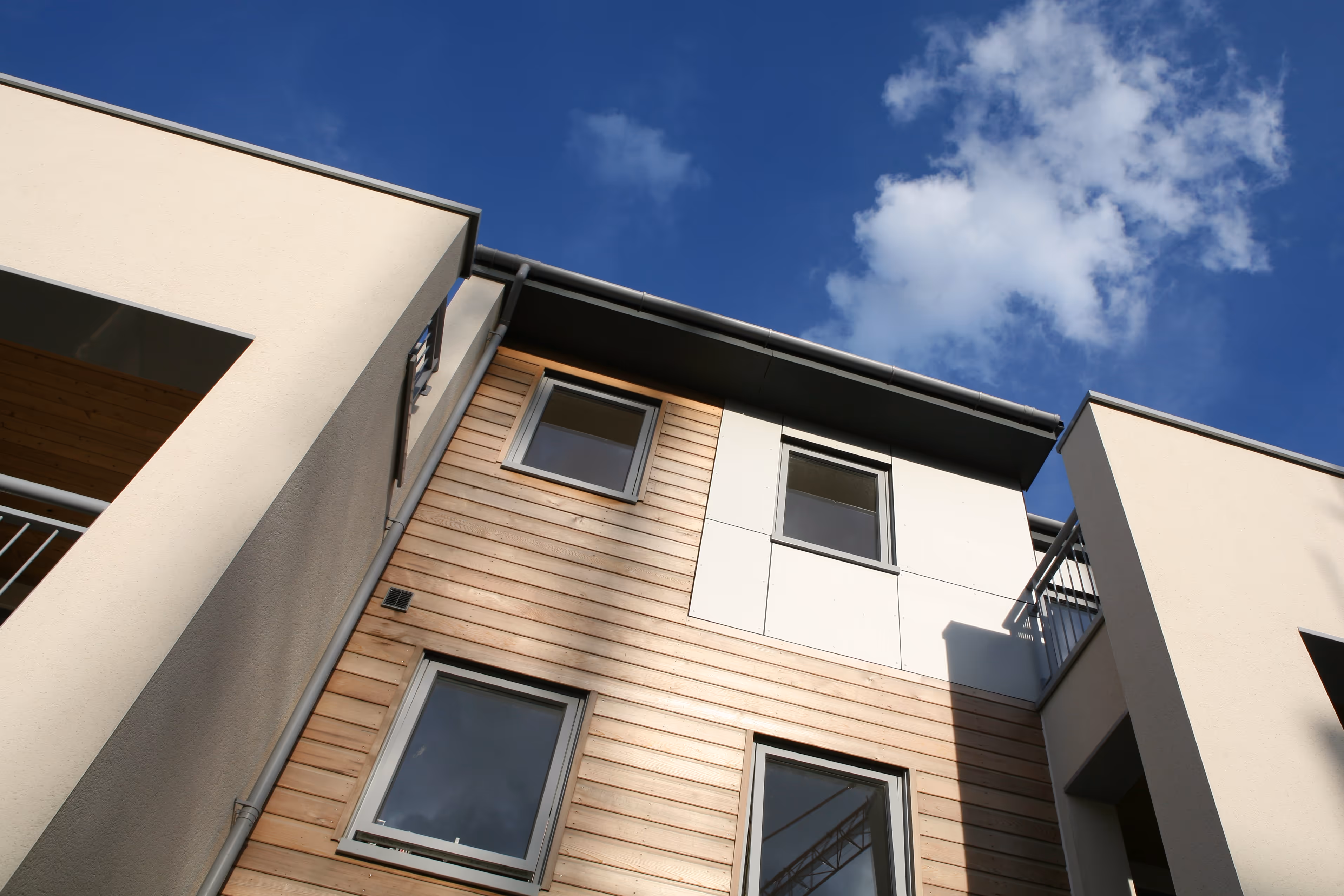 Modern building facade with wooden and white paneling, featuring four windows under a blue sky with a few clouds.