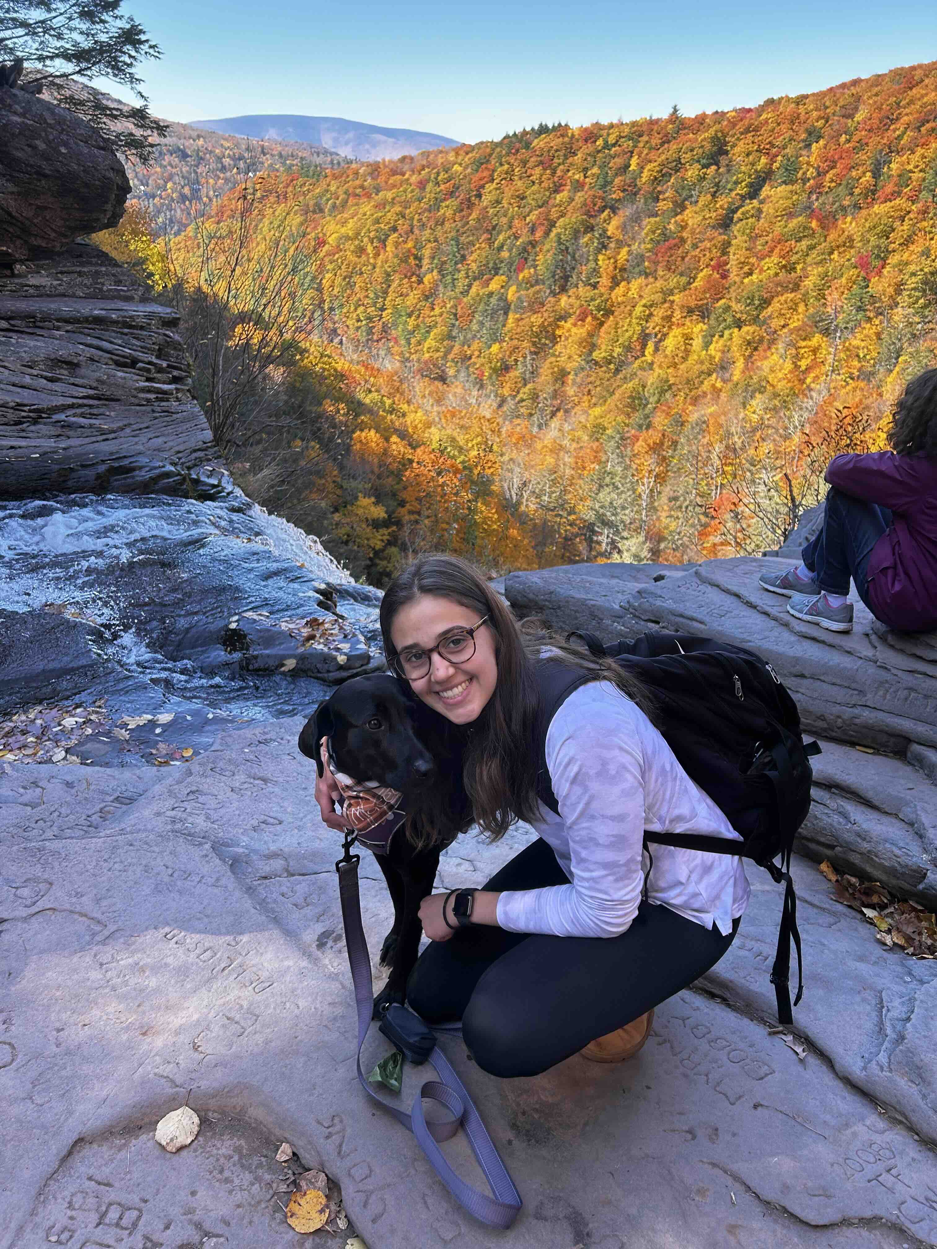 Young woman with glasses and a backpack crouching and hugging a black Labrador on a rocky overlook with autumn-colored forest in the background.