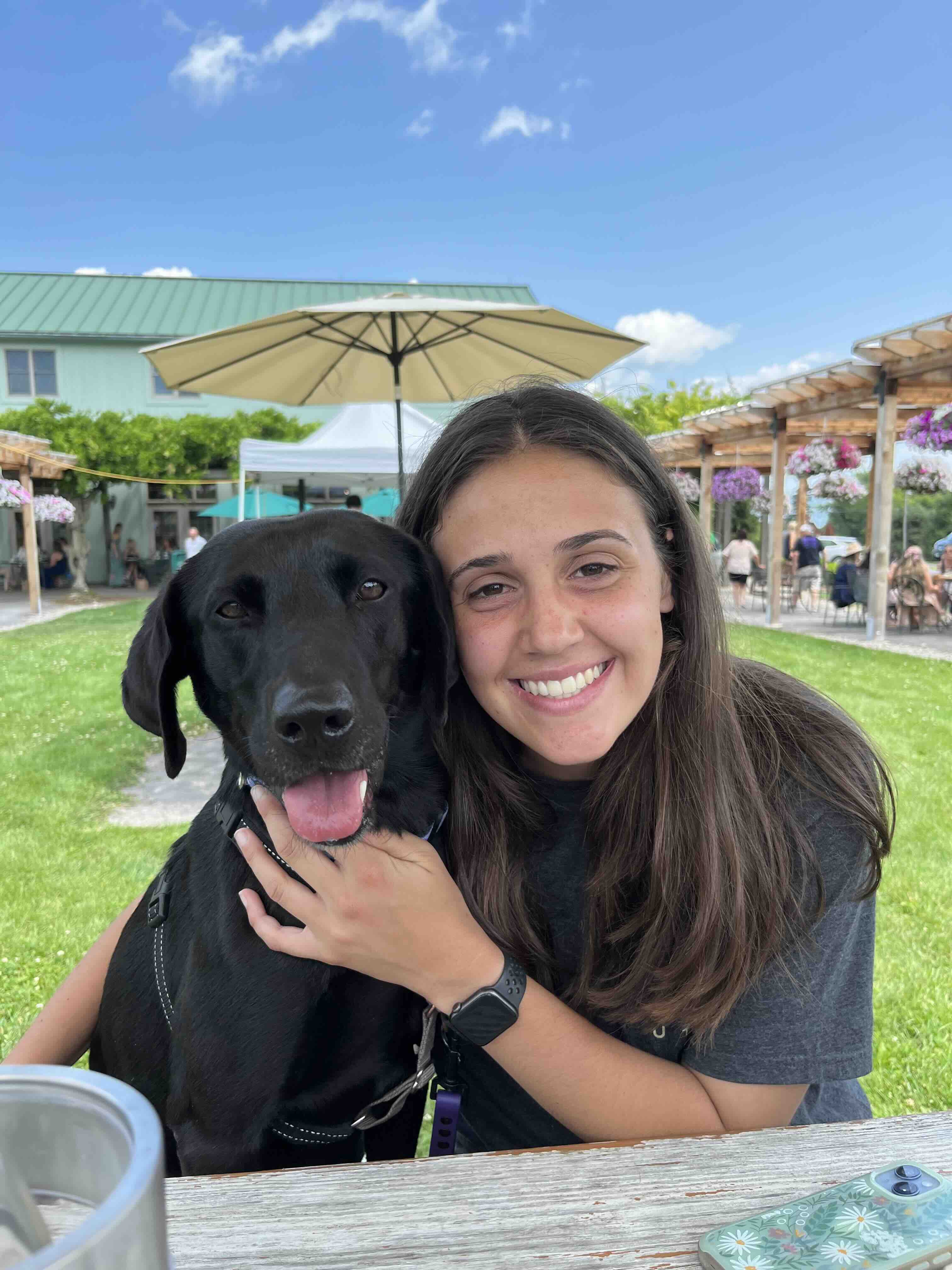 Smiling woman hugging a happy black Labrador retriever sitting outdoors at a picnic table.