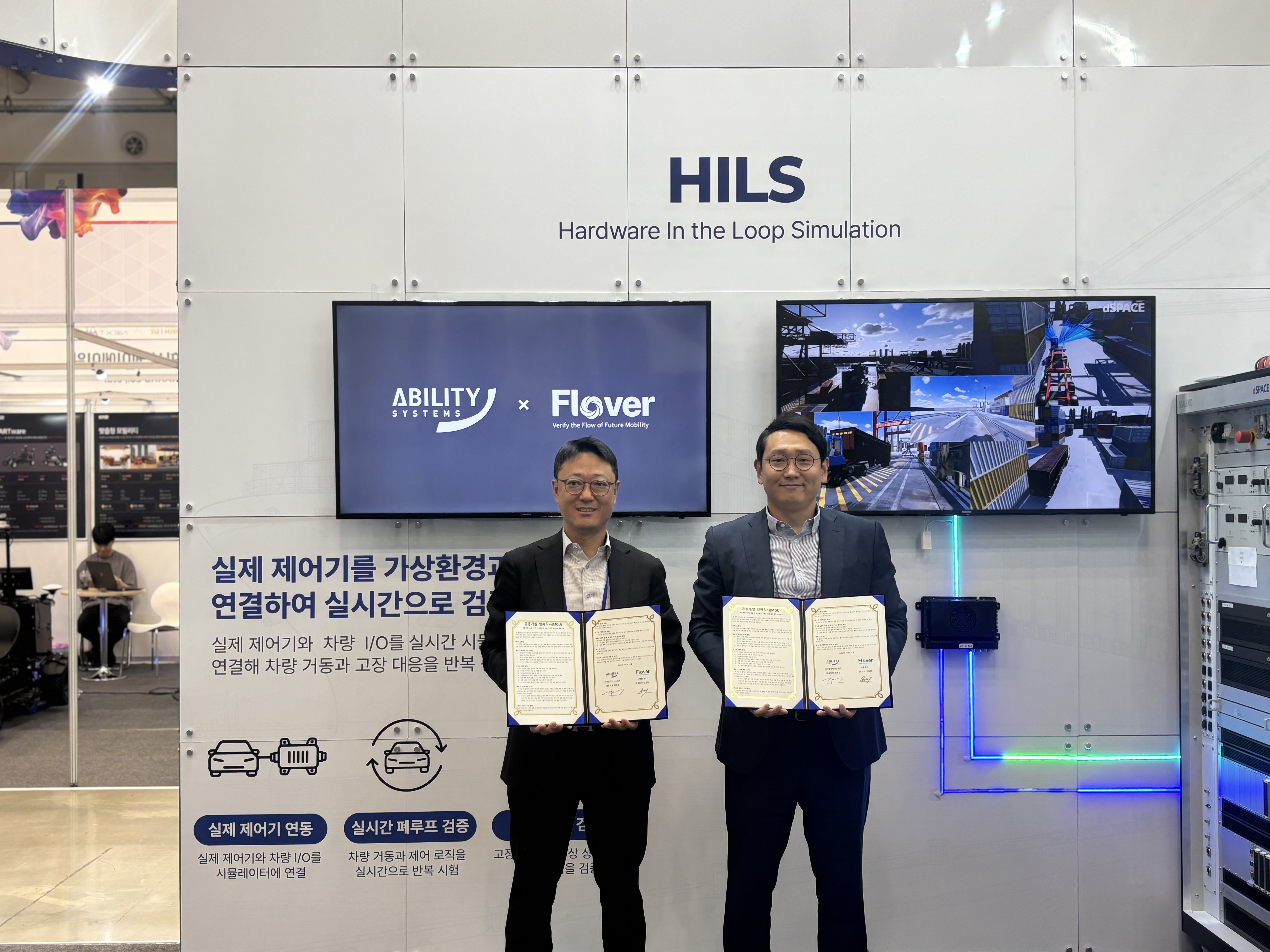 임동현 플로버 대표와 신재일 어빌리티시스템즈 대표가 ABILITY SYSTEMS × Flover 화면과 HILS 검증 장비 앞에서 업무협약서를 들고 있는 모습