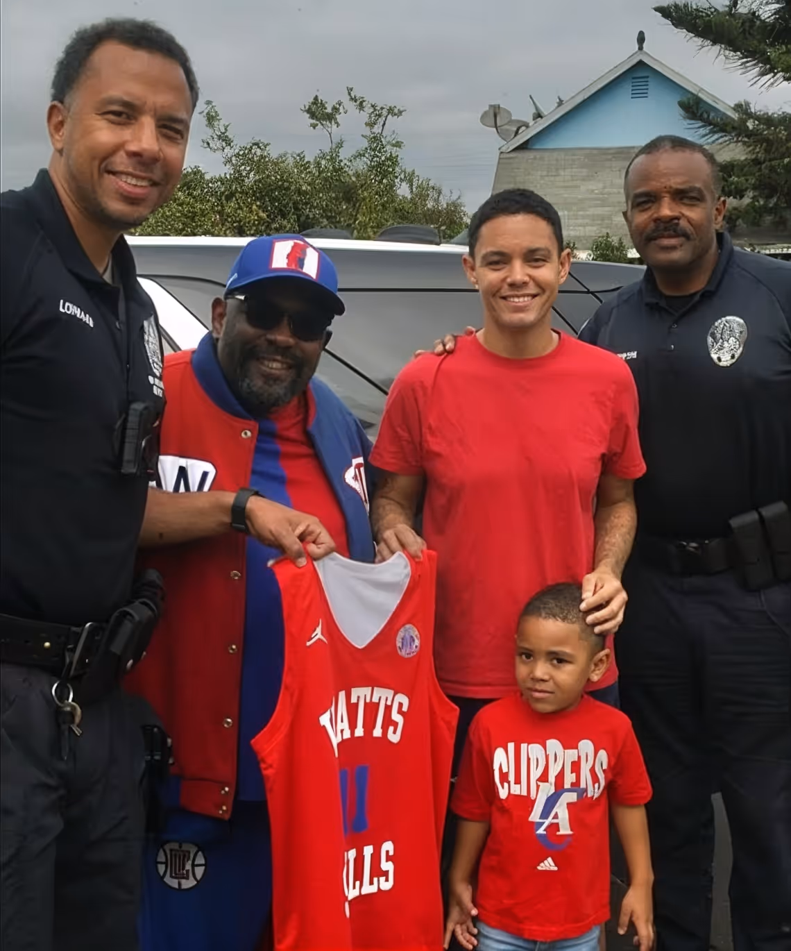 Two police officers and three people, including a child, smiling outdoors with one person holding a red Watts Hills basketball jersey.