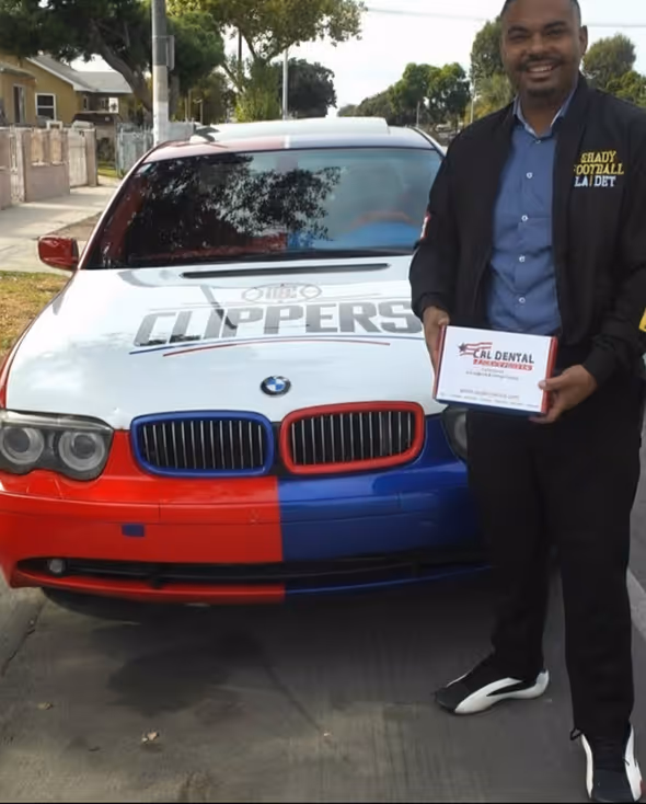 Man standing next to a BMW car painted in red, white, and blue colors with 'Clippers' logo on hood, holding a Cal Dental Care Services box.