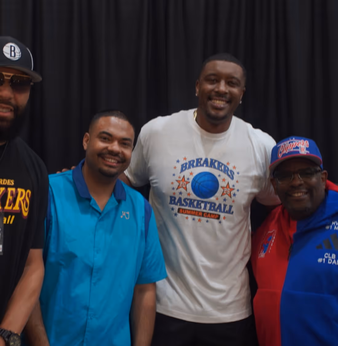 Four men smiling and posing in front of a black curtain, with one wearing a white 'Breakers Basketball Summer Camp' shirt.