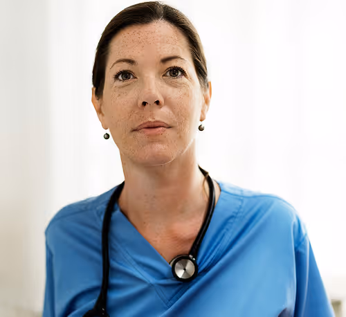 Female healthcare professional in blue scrubs with a stethoscope around her neck looking slightly upwards.