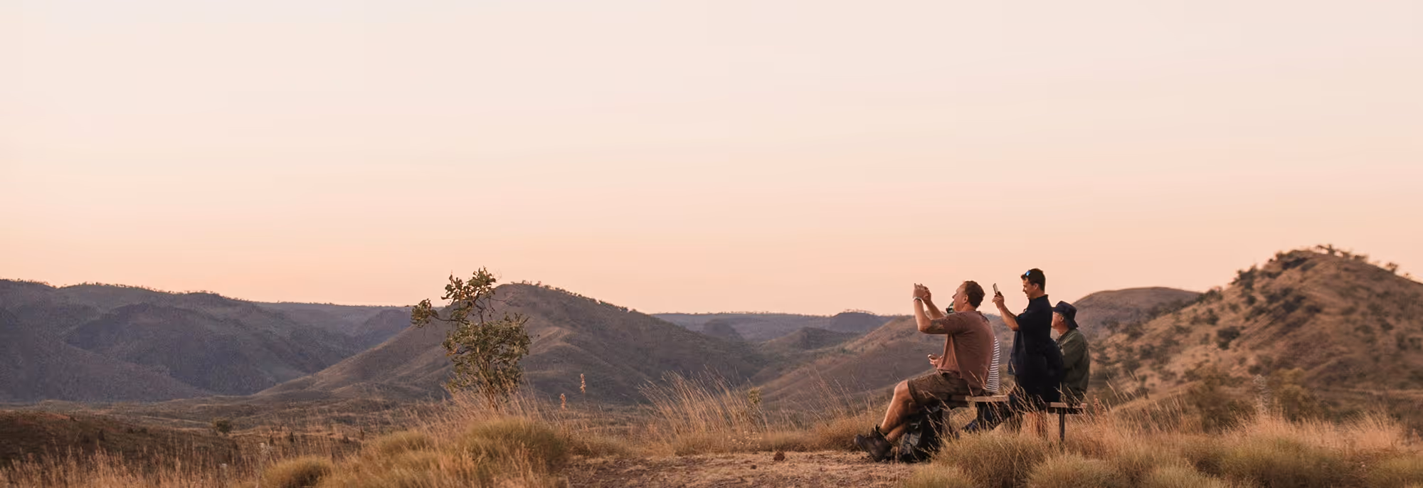Three people sitting and standing on a bench in a dry grassy landscape with hills in the background at sunset, taking photos with their phones.
