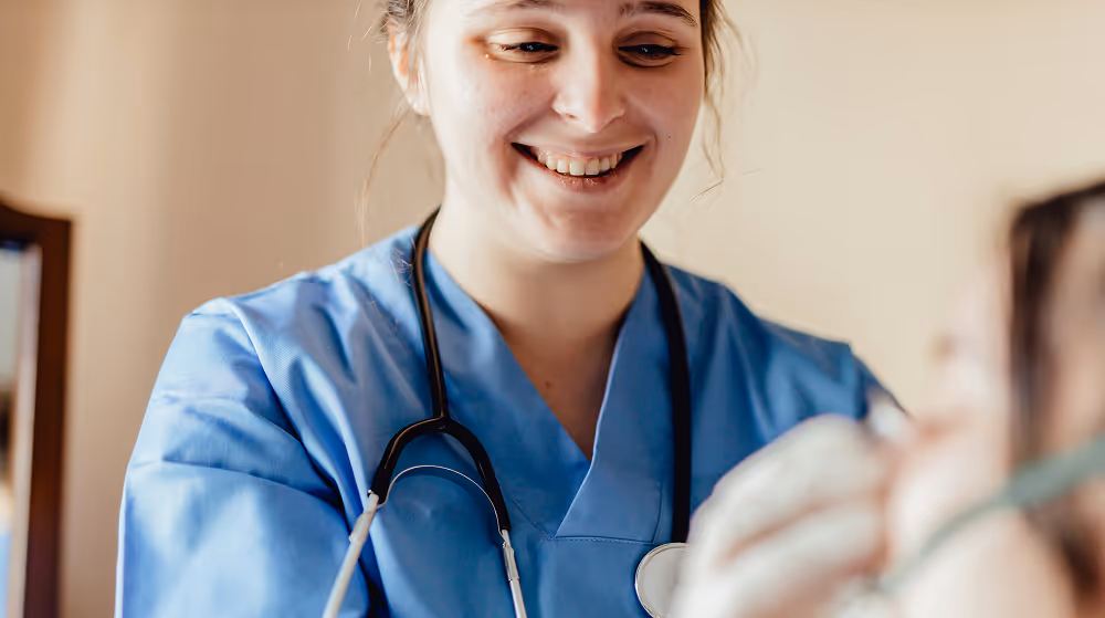 Smiling nurse in blue scrubs with stethoscope attending to a patient.