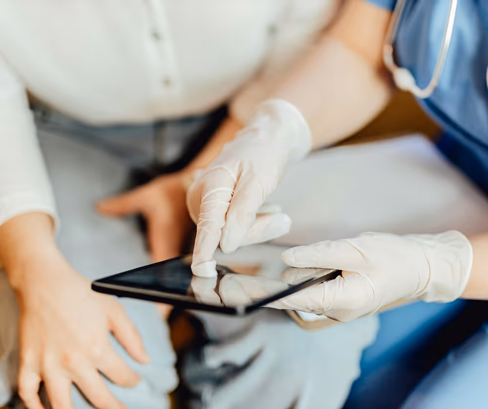Healthcare professional wearing gloves using a tablet while consulting with a patient.
