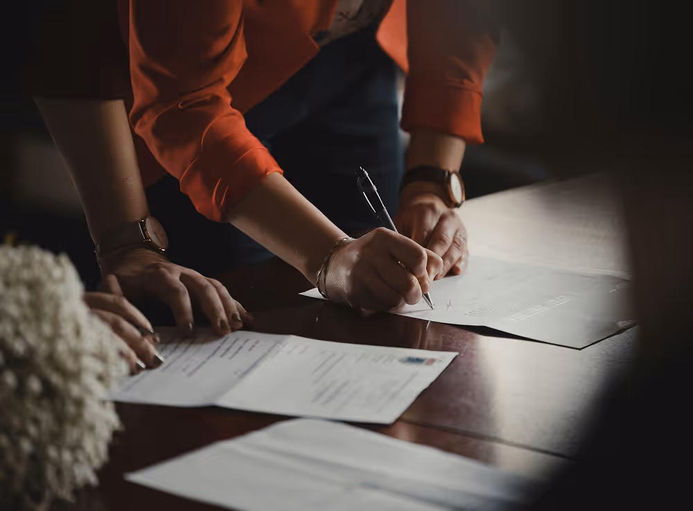 Person in an orange shirt signing a document on a wooden table with other papers nearby.
