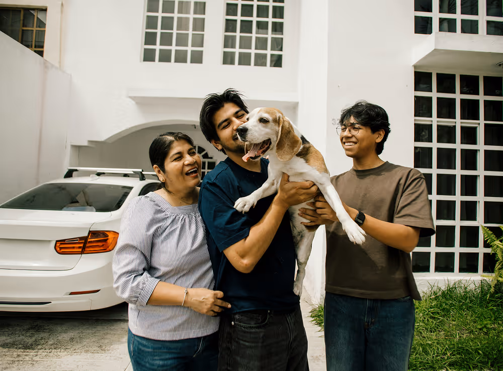Three people smiling and holding a happy beagle dog outside a house with a white car parked nearby.