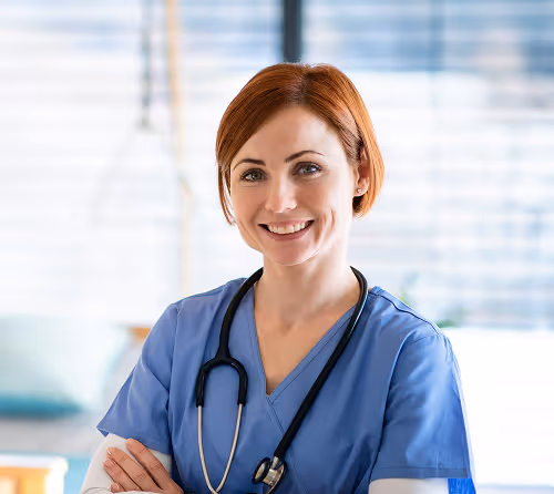 Smiling female healthcare professional wearing blue scrubs and a stethoscope in a bright medical office.