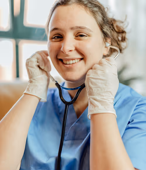 Smiling female healthcare worker in blue scrubs holding a stethoscope to her ears while wearing gloves.