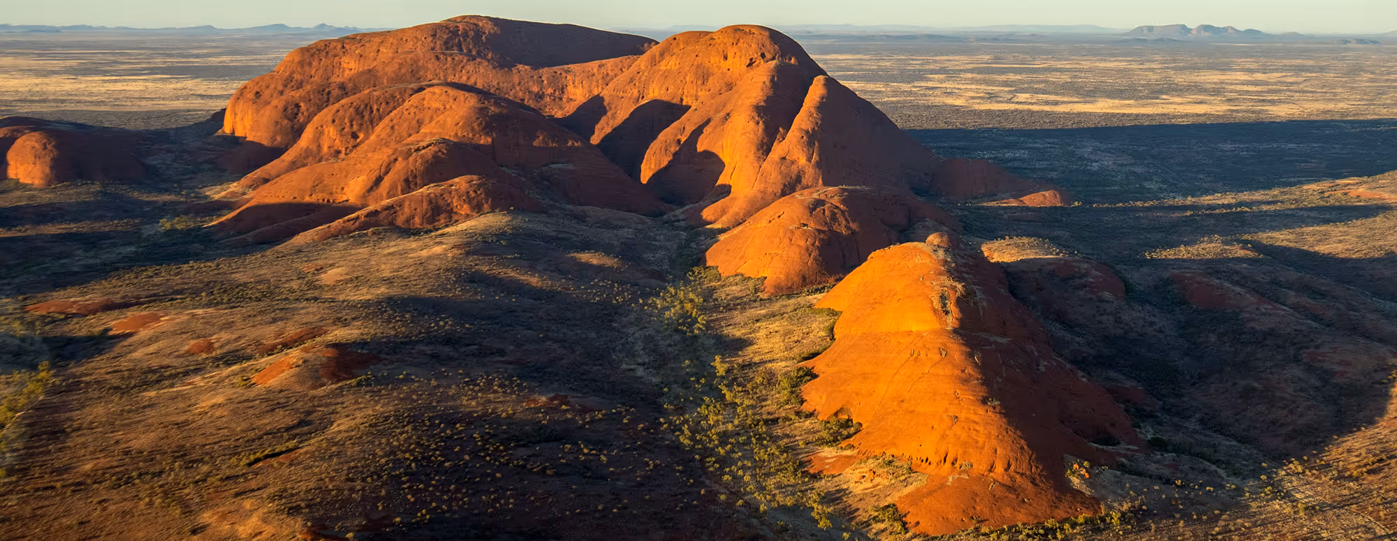 Sunlit red rock formations with scattered vegetation in a vast arid landscape during late afternoon.