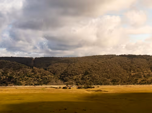 Wide landscape showing green fields with dense forested hills under a cloudy sky.