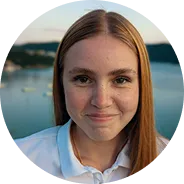 Smiling young woman with straight light brown hair and freckles, wearing a white collared shirt, with a blurred waterfront background.