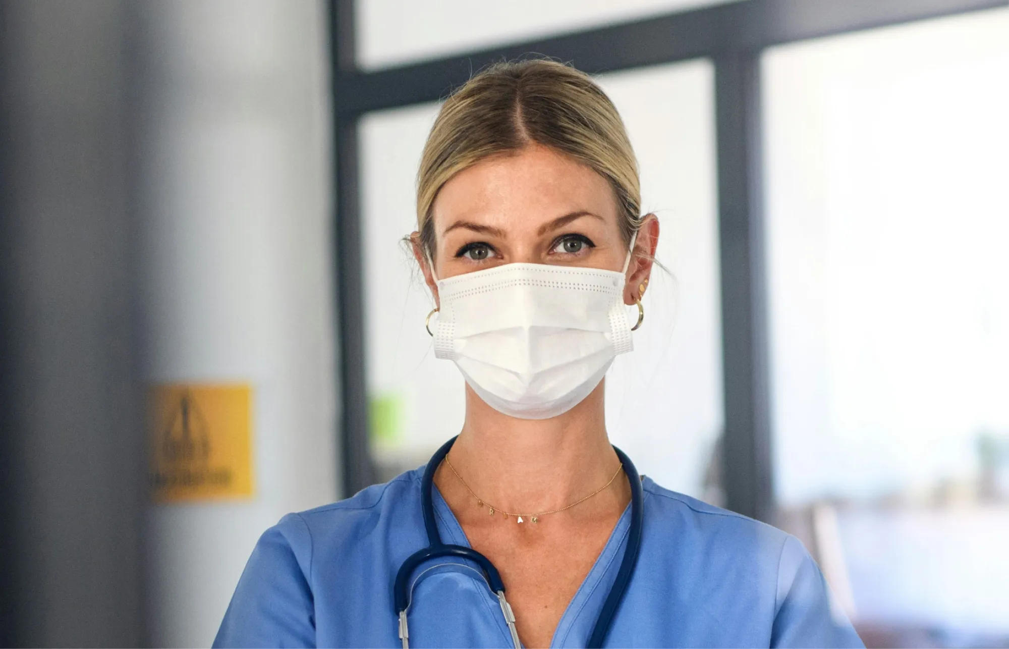 Female healthcare professional wearing a surgical mask and blue scrubs with a stethoscope around her neck.