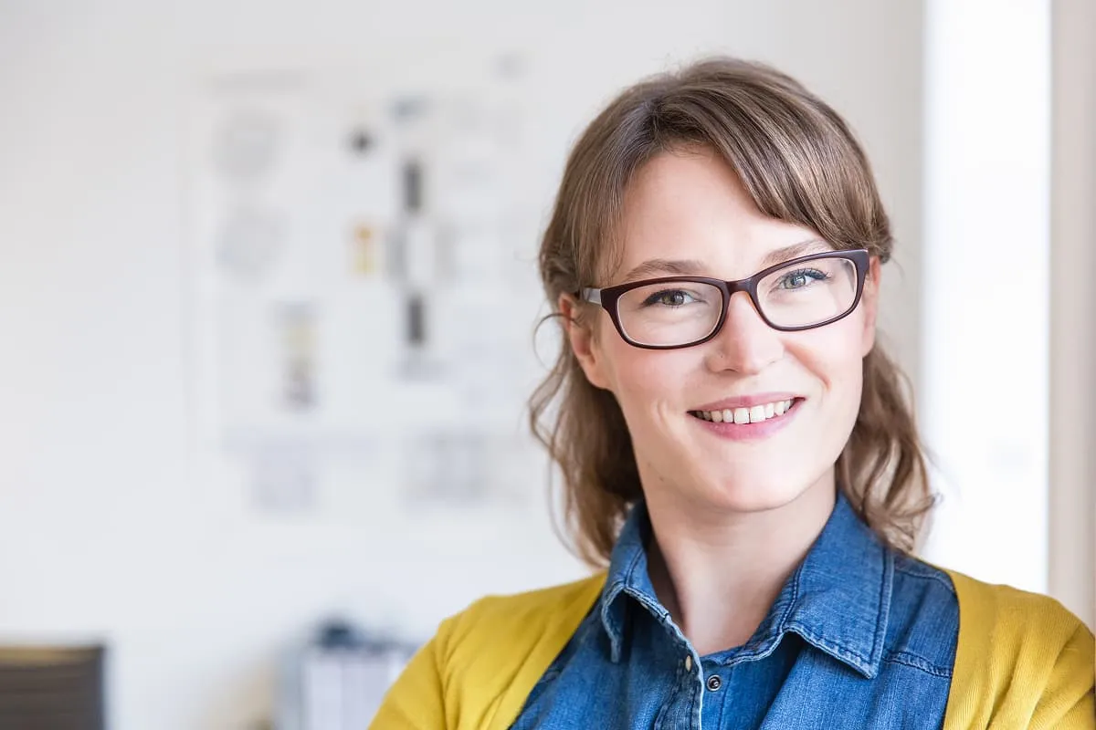 Smiling woman wearing glasses, a denim shirt, and a yellow cardigan in a bright room.
