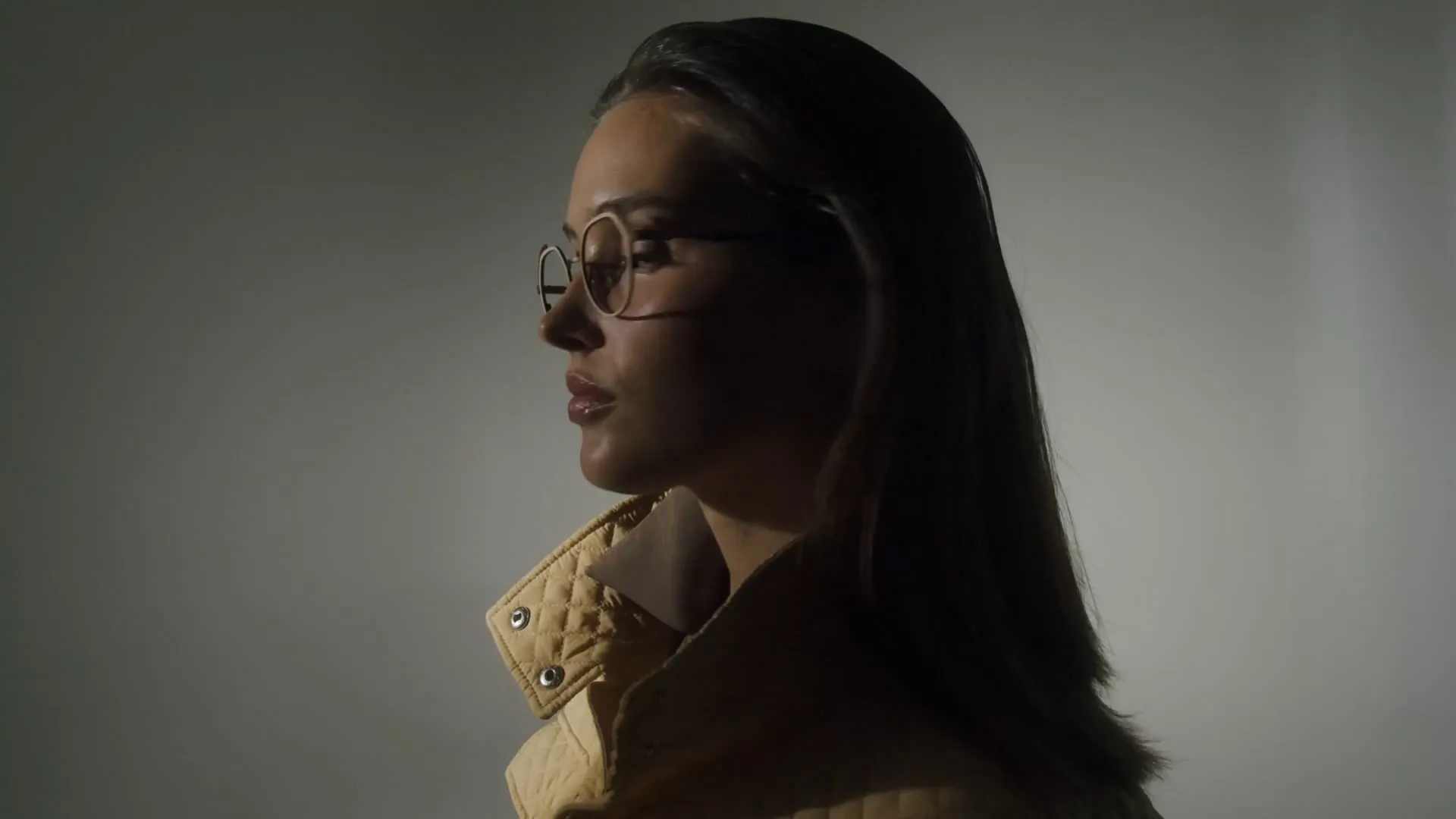Side profile of woman with glasses and long hair wearing a quilted beige jacket against gray background.