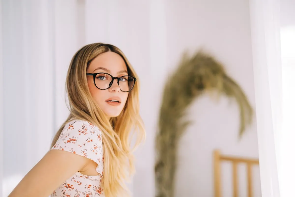 Blonde woman with glasses wearing a floral short-sleeve top, looking at the camera indoors.