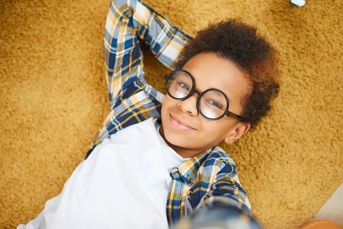 Smiling boy wearing glasses and plaid shirt lying on tan carpet taking a selfie.