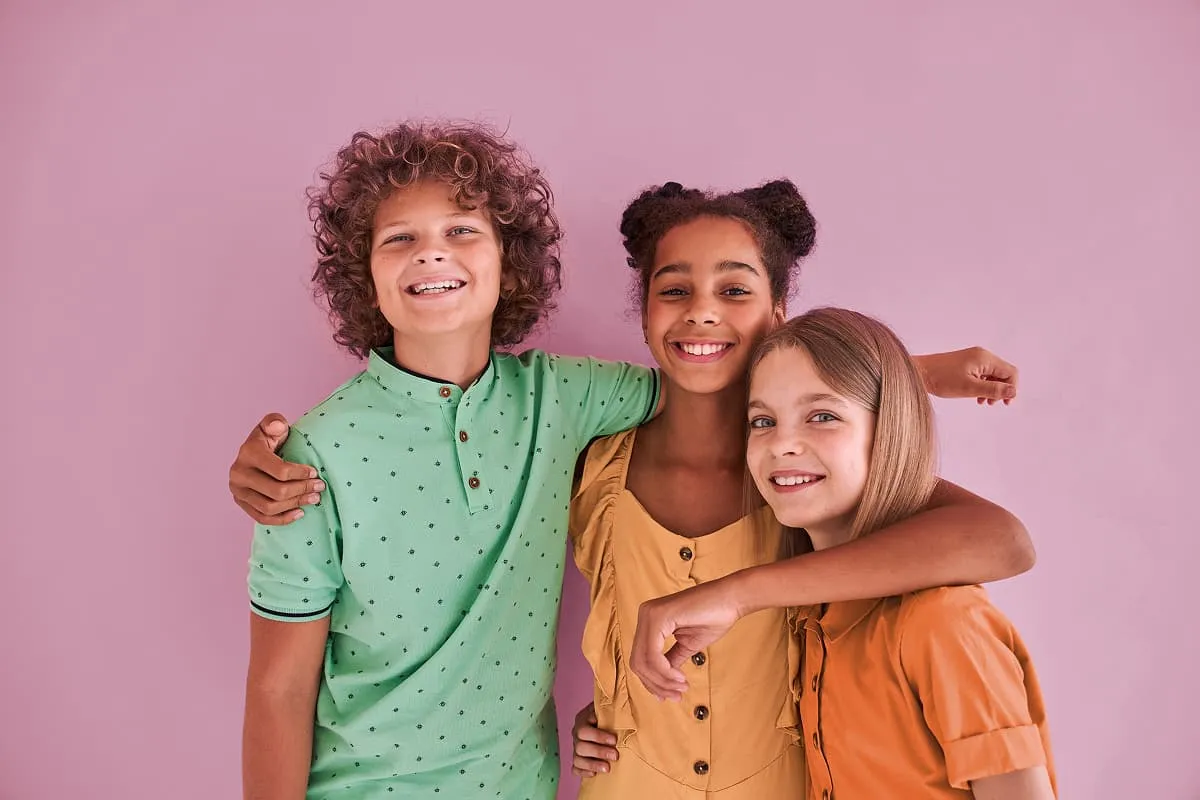 Three smiling children standing close with arms around each other against a pink background.
