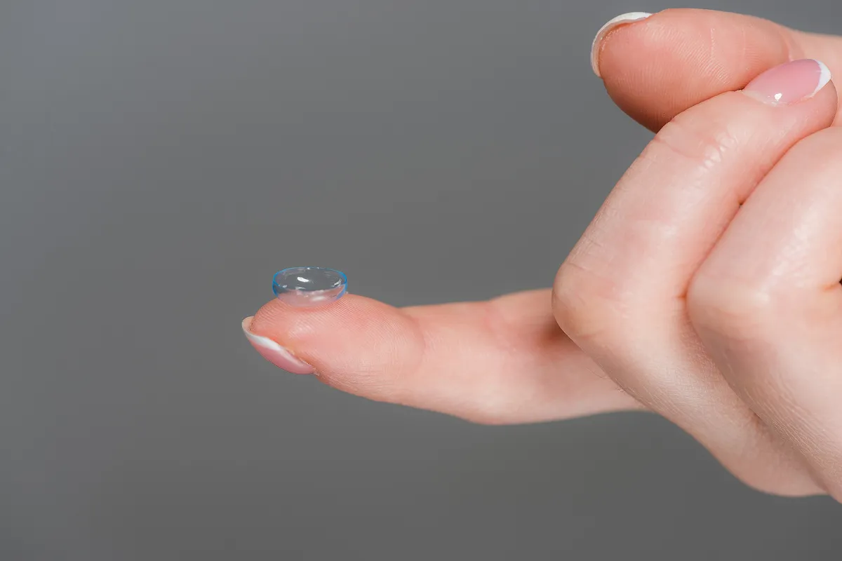 Hand holding a transparent contact lens balanced on the tip of an index finger against a gray background.