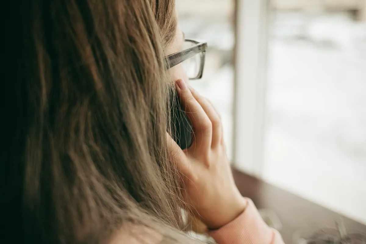 Woman wearing glasses holds phone to ear near window with blurred background.