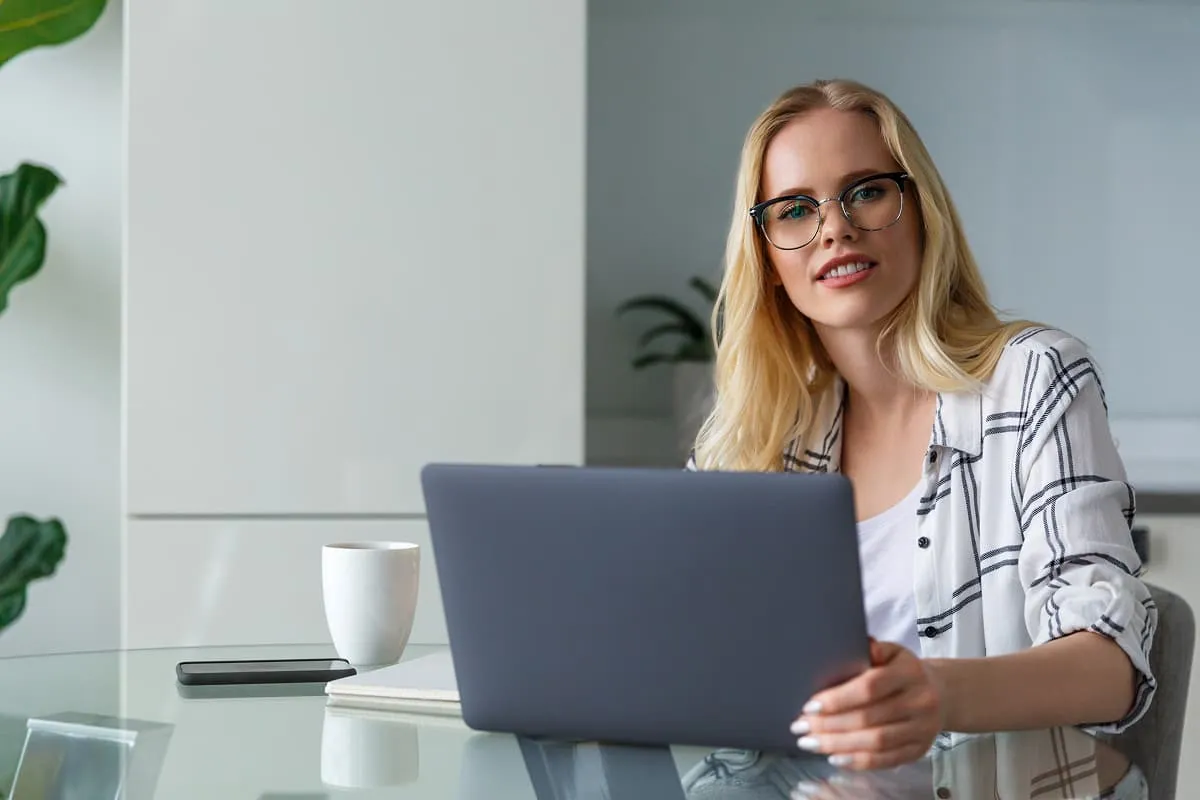 Young woman with glasses seated at glass table holding laptop, smiling toward camera.
