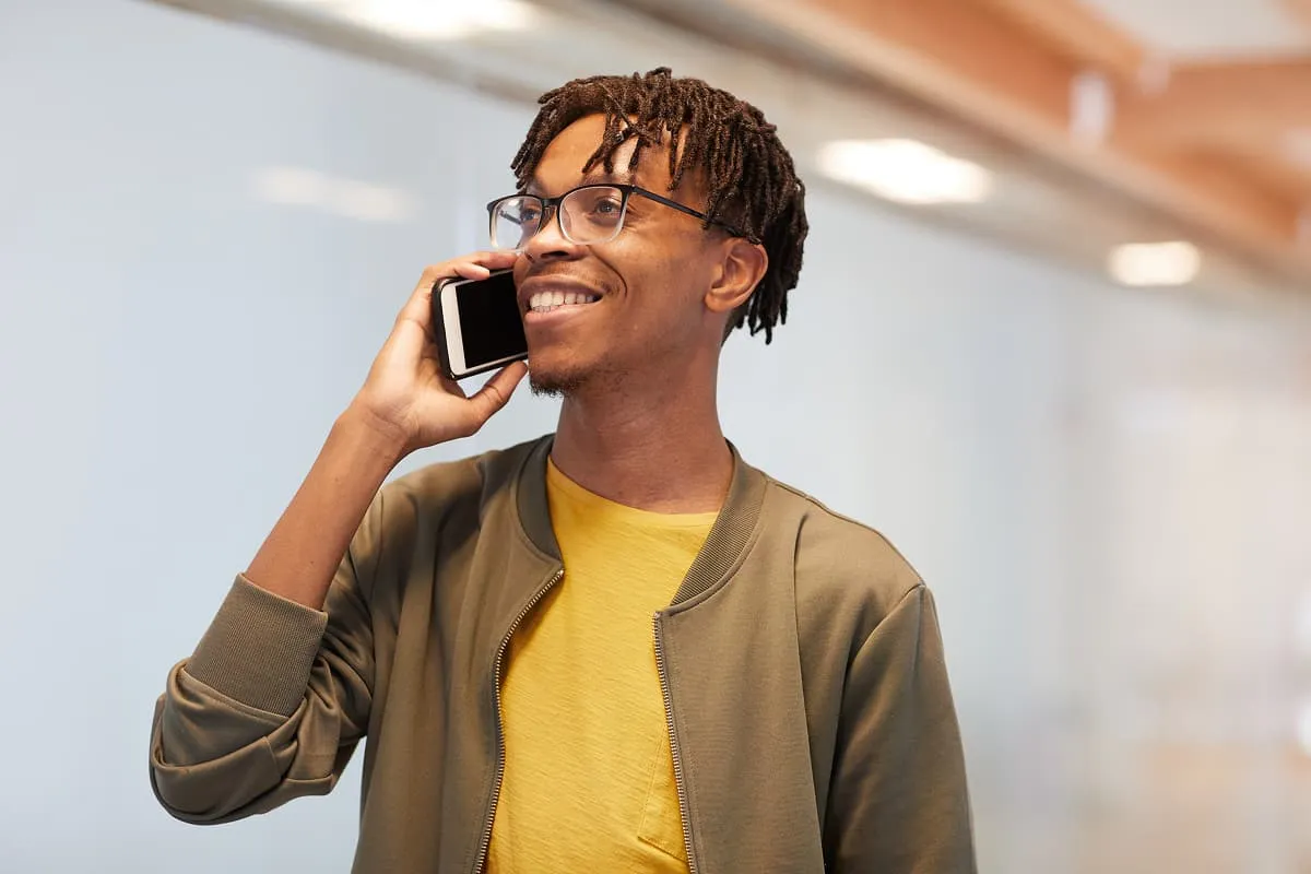 Smiling young man with glasses talking on a smartphone, wearing a yellow shirt and olive jacket.
