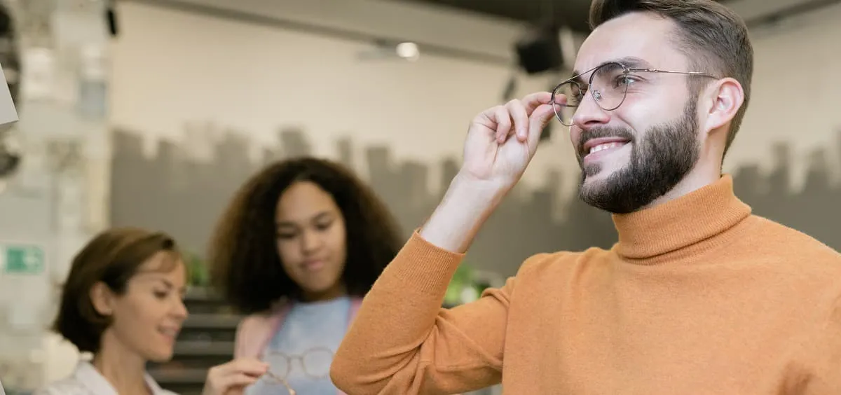 Young man with beard smiling while trying on eyeglasses in a store with two women in background.