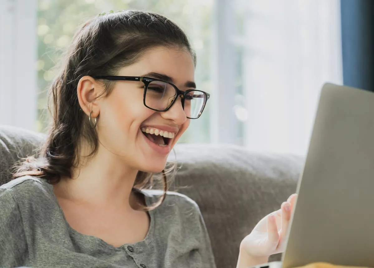 Young woman with glasses laughing while looking at a laptop.