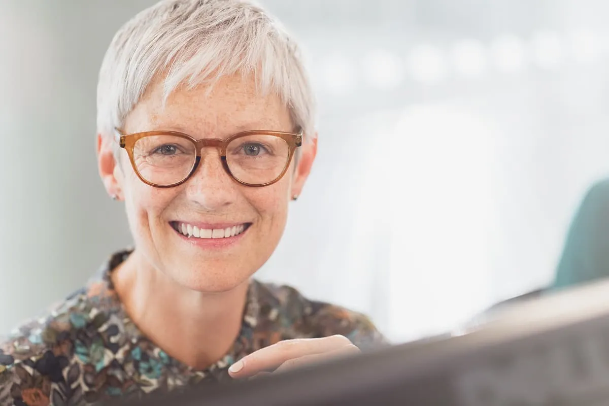Smiling senior woman with short white hair and glasses wearing a floral blouse indoors.