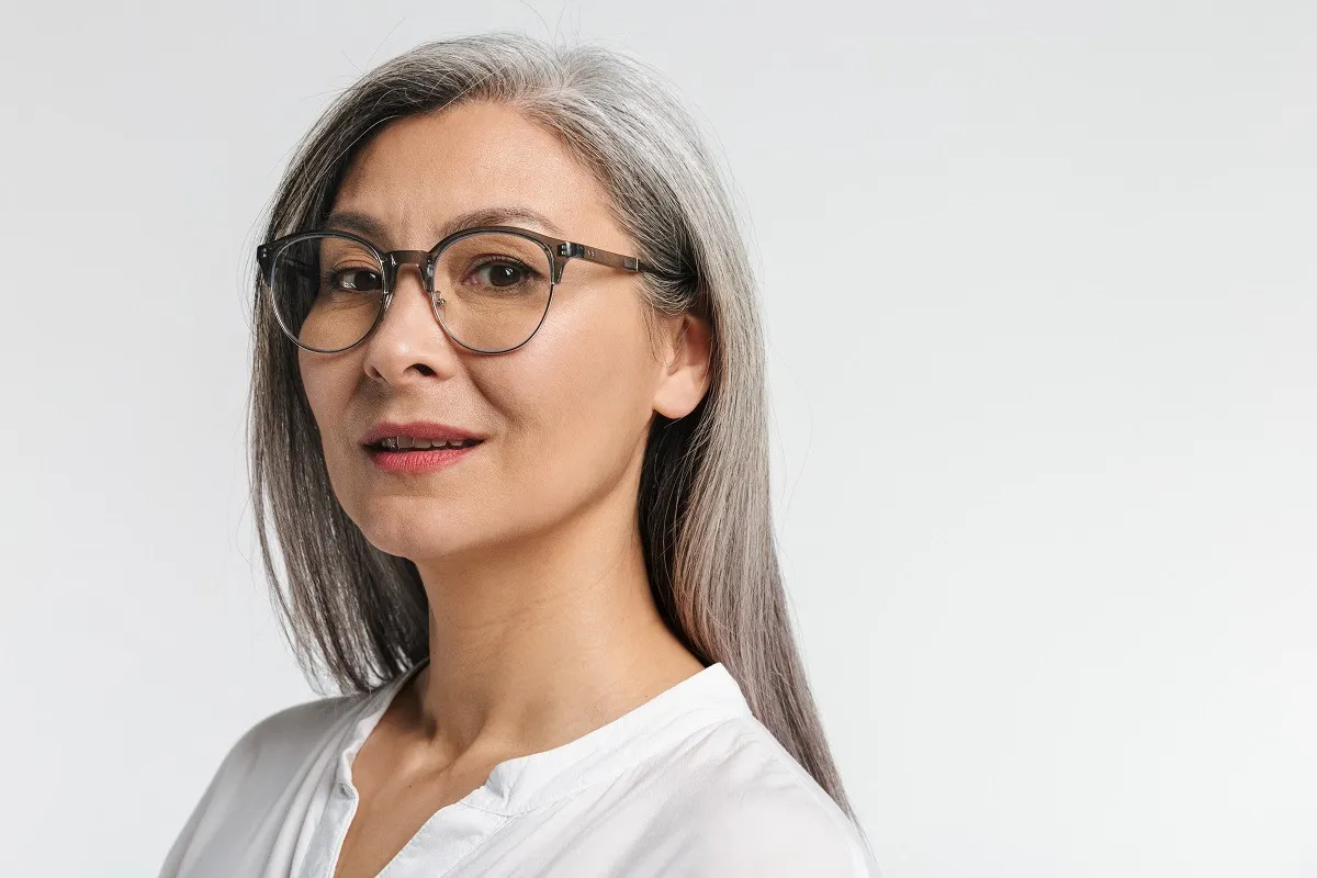 Smiling middle-aged woman with gray hair and glasses wearing a white top on a light background.