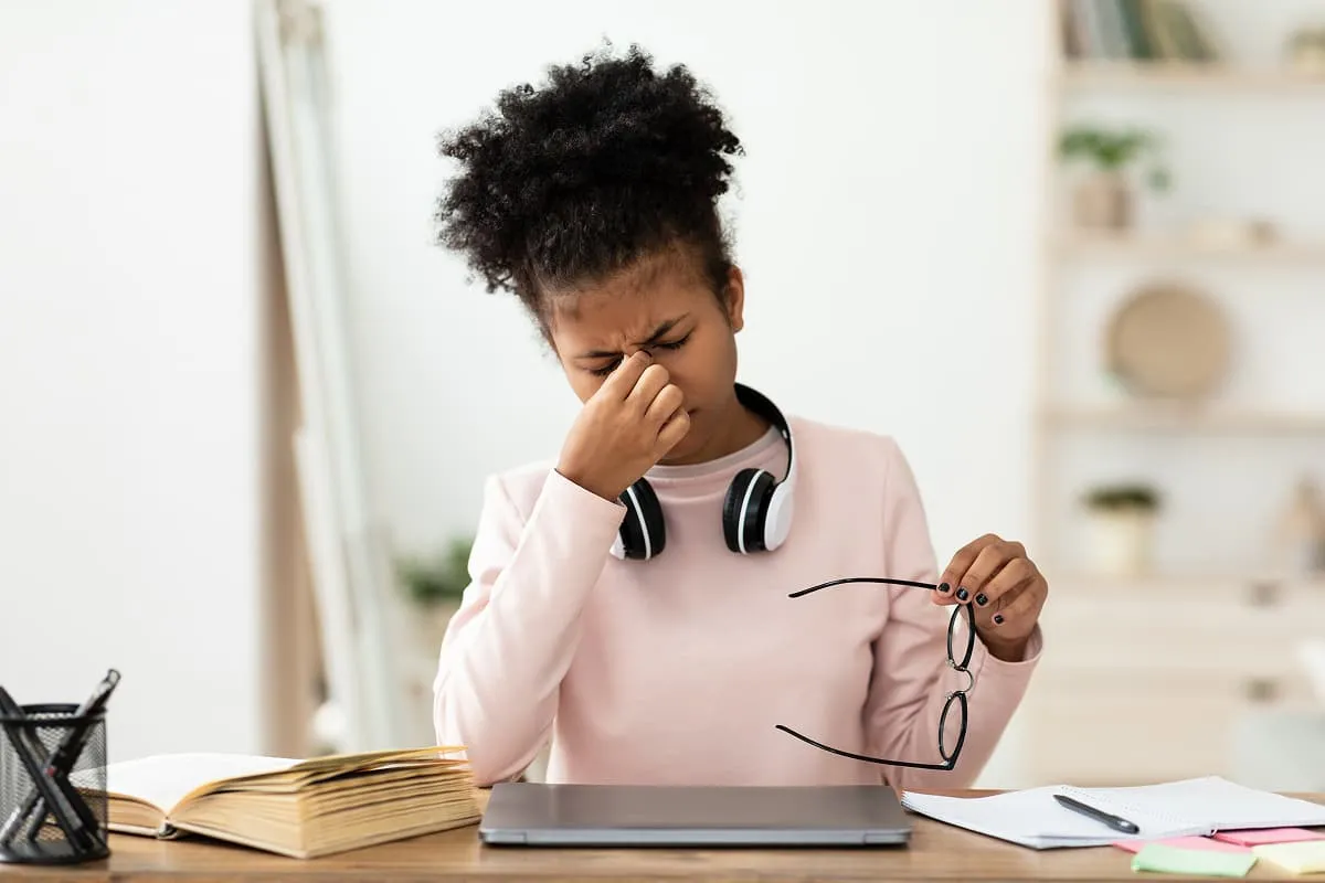 Young woman at desk rubbing eyes holding glasses with laptop, open book, and papers around her.