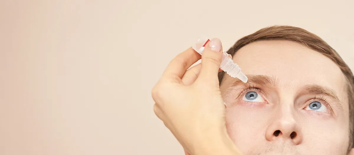 Close-up of a man applying eye drops to his right eye against a beige background.
