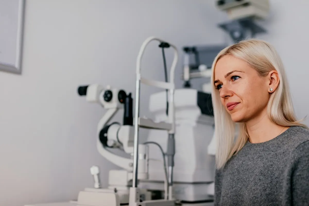 Blonde woman sitting beside an optometry machine in an eye exam room.