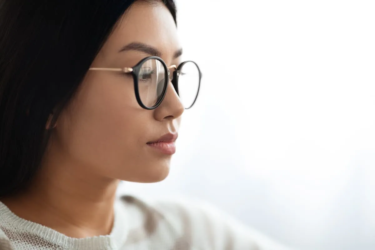 Side profile of a young woman wearing round glasses and a light-colored sweater against a plain background.