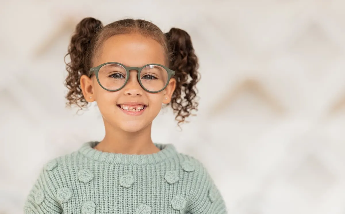 Smiling young girl with glasses and curly pigtails wearing a light green sweater.